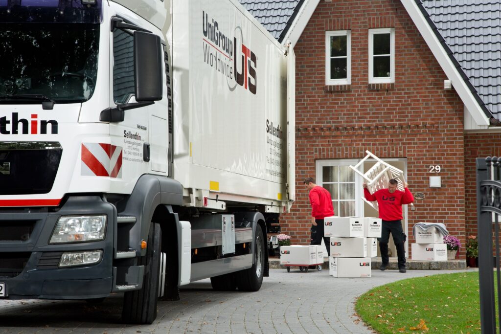 Ein Umzugswagen mit der Aufschrift „UniGroup Worldwide“ steht vor einem Haus. Zwei Umzugshelfer in roten Hemden tragen Kisten zum Eingang.