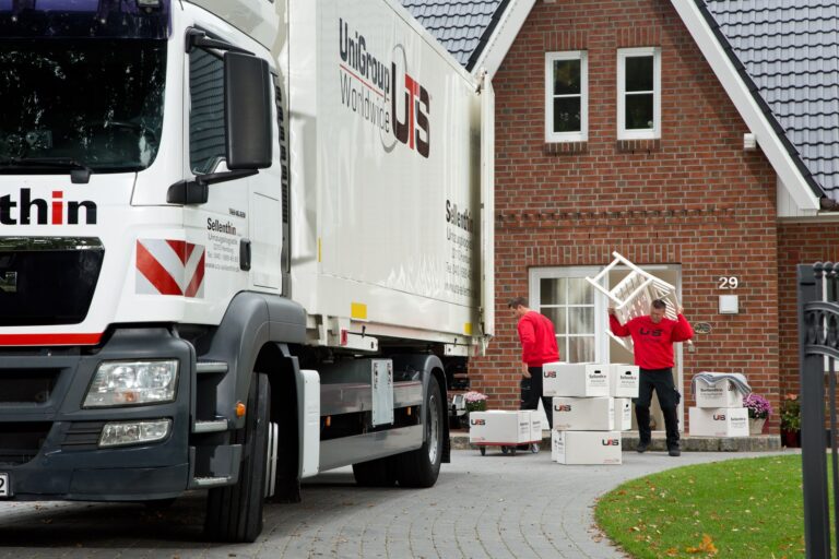 Ein Umzugswagen mit der Aufschrift „UniGroup Worldwide“ steht vor einem Haus. Zwei Umzugshelfer in roten Hemden tragen Kisten zum Eingang.