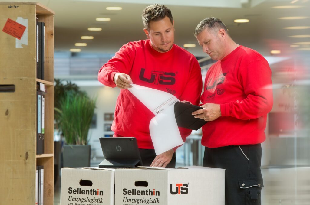 Zwei Männer in roten UTS-Pullovern arbeiten gemeinsam in einem hellen Raum und bedienen elektronische Geräte in der Nähe von beschrifteten Sellenthin-Umzugskartons.