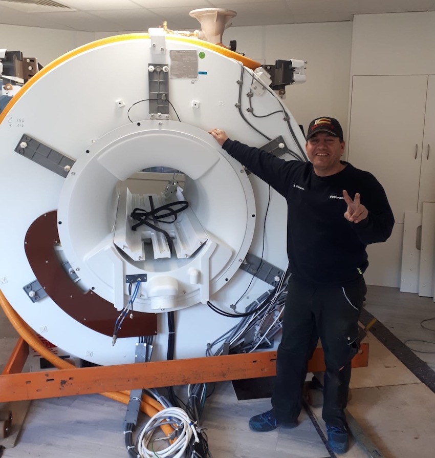 Ein Mann steht neben einem großen MRT-Gerät in einem Raum. Das Gerät ist weiß und hat sichtbare Kabel und Metallteile.