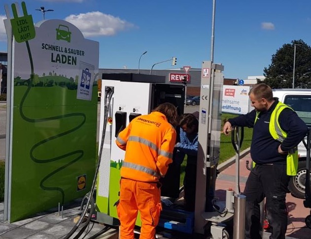 An einem sonnigen Tag arbeiten drei Personen an einer Ladestation für Elektrofahrzeuge.