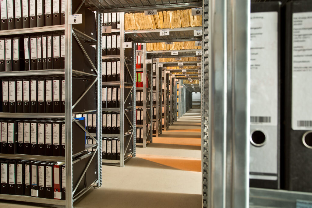 Lange Reihen von Metallregalen, gefüllt mit ordentlich angeordneten schwarzen Ordnern und beigen Mappen, in einem gut beleuchteten Archivraum.