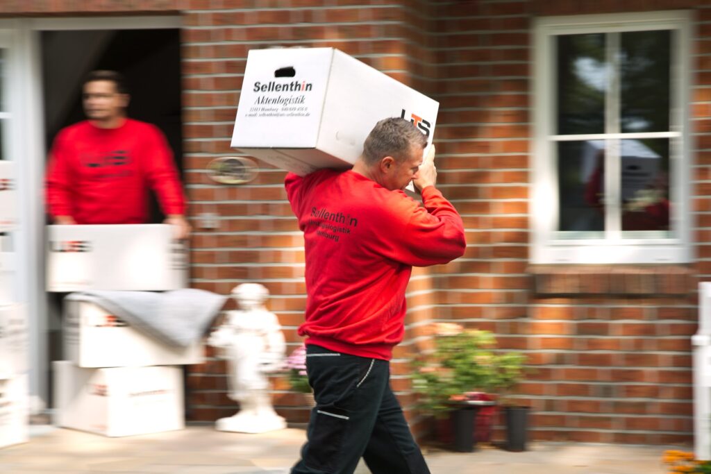 Ein Mann in einem roten Sweatshirt trägt eine große Kiste mit der Aufschrift „Sellenthin Aktenlogistik“ aus einem Gebäude.