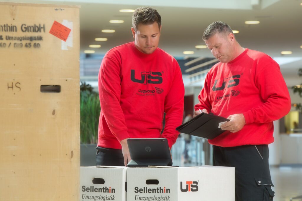 Zwei Männer in roten UTS-Pullovern arbeiten gemeinsam in einem hellen Raum und bedienen elektronische Geräte in der Nähe von beschrifteten Sellenthin-Umzugskartons.