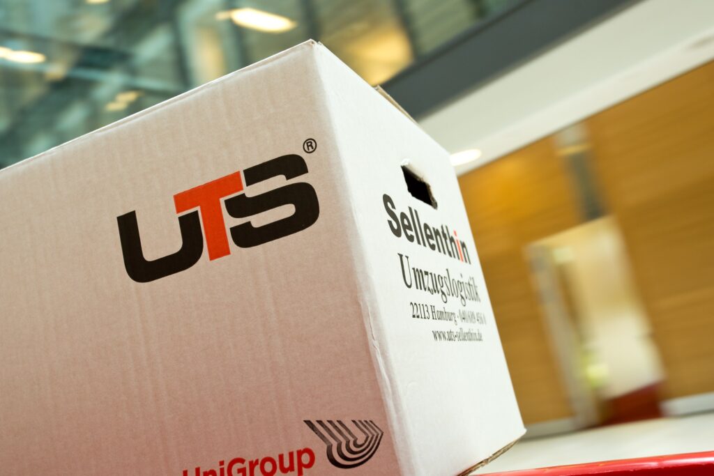 Umzugskarton mit „UTS“-Logo und dem Schriftzug „Sellenthin Umzugslogistik“, positioniert in einer modernen Büroumgebung.