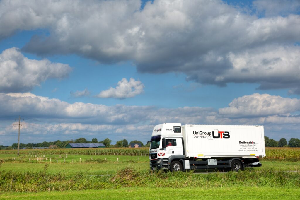 Ein Lkw mit dem Logo „UniGroup Worldwide UTS“ und "Sellenthin" fährt unter einem teilweise bewölkten blauen Himmel durch eine grün bewachsene Wiese.