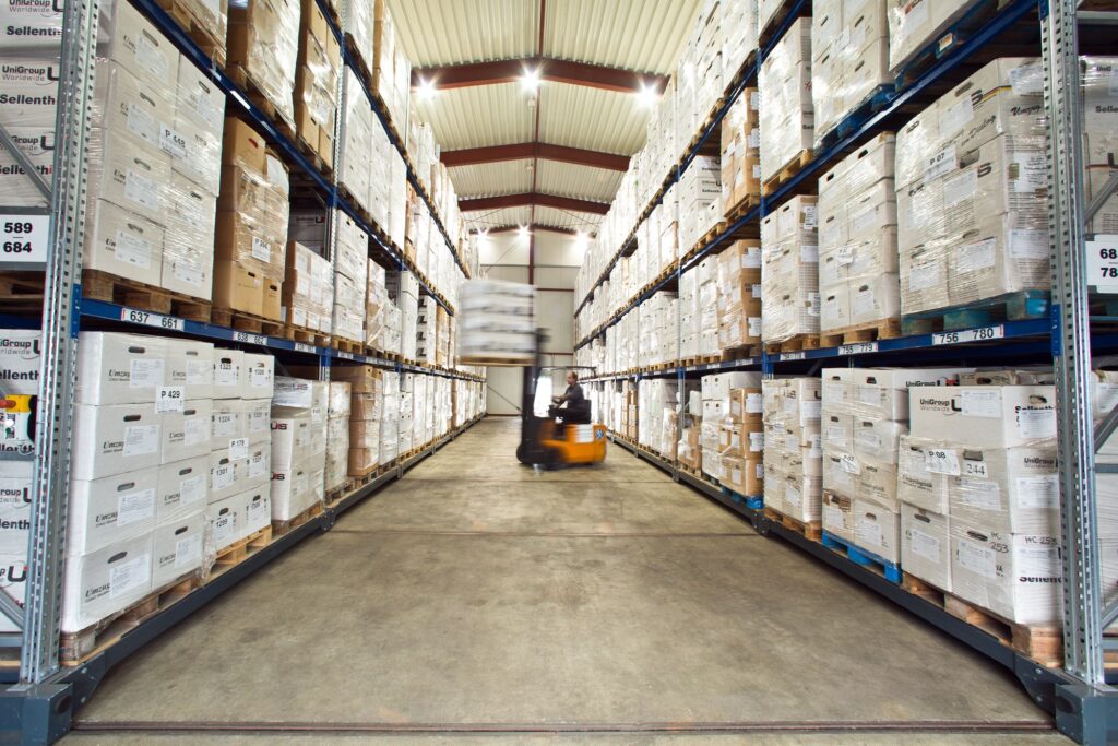 Breiter Gang in einem gut beleuchteten Lagerhaus mit hohen Regalen, die mit Kartons bestückt sind. Ein Gabelstapler mit verschwommener Bewegung transportiert Gegenstände.