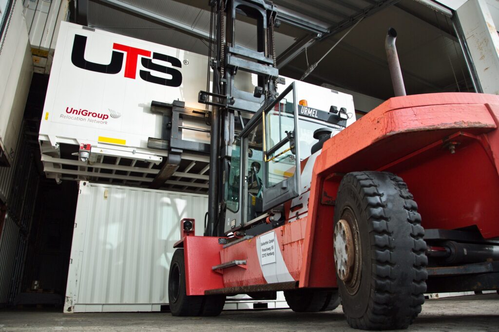Ein Gabelstapler hebt einen großen Container mit der Aufschrift „UTS UniGroup Relocation Network“ in einem Lagerhaus an.