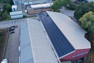 Luftbild eines großen Lagerhauses mit Solarzellen auf dem Dach, umgeben von Bäumen und Straßen.