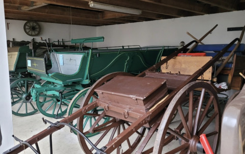 Eine Sammlung von Oldtimer-Kutschen in einer schwach beleuchteten Garage. Im Vordergrund steht ein brauner Holzkarren mit Griffen, im Hintergrund eine grüne Kutsche.