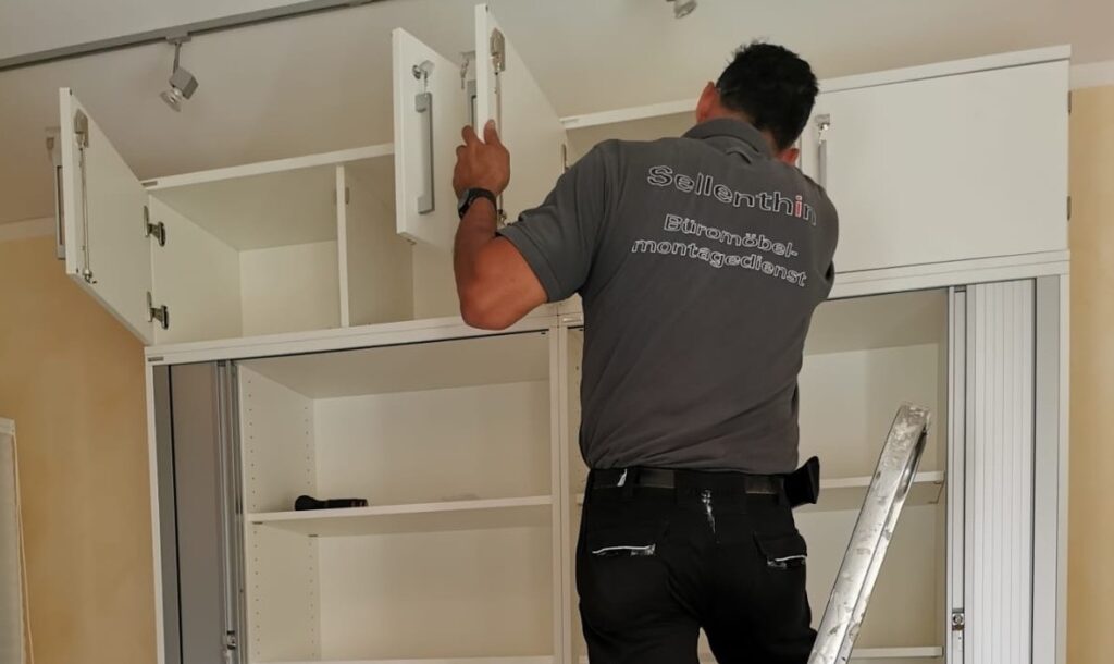 Ein Mann steht auf einer Leiter und installiert oder inspiziert offene weiße Schränke an der Wand.