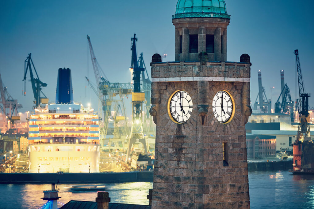 Der Uhrturm mit beleuchteten Zifferblättern steht an einem Hafen mit einem hell erleuchteten Kreuzfahrtschiff und Kränen im Hintergrund.
