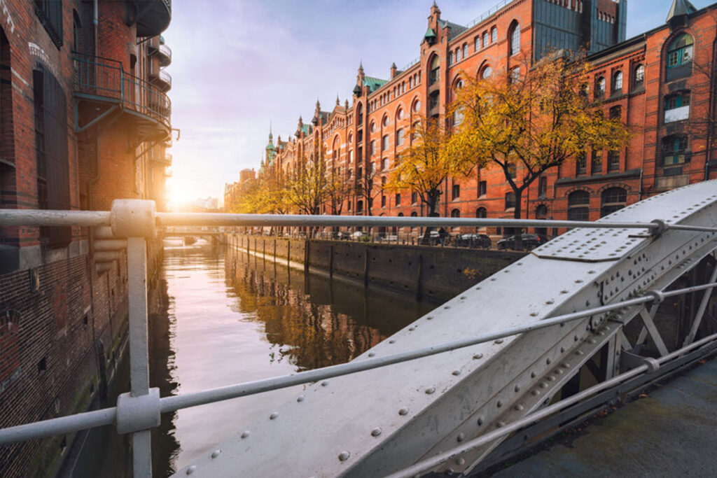 Sonnendurchfluteter Blick auf ein Lagerhausviertel aus rotem Backstein mit einem ruhigen Kanal und Bäumen mit Herbstlaub.