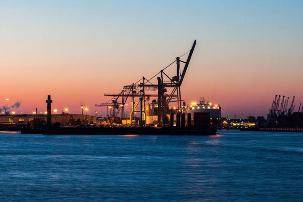 Ein Sonnenuntergang über einem geschäftigen Hafen, dessen Kräne sich vor einem leuchtend orange-rosa Himmel abzeichnen.