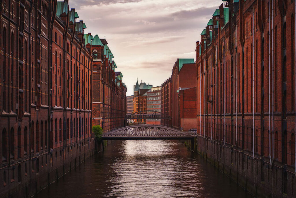 Eine ruhige Kanalszene bei Sonnenuntergang, flankiert von historischen roten Backsteinlagern mit grünen Dächern und einer Brücke, die sich über das Wasser spannt.