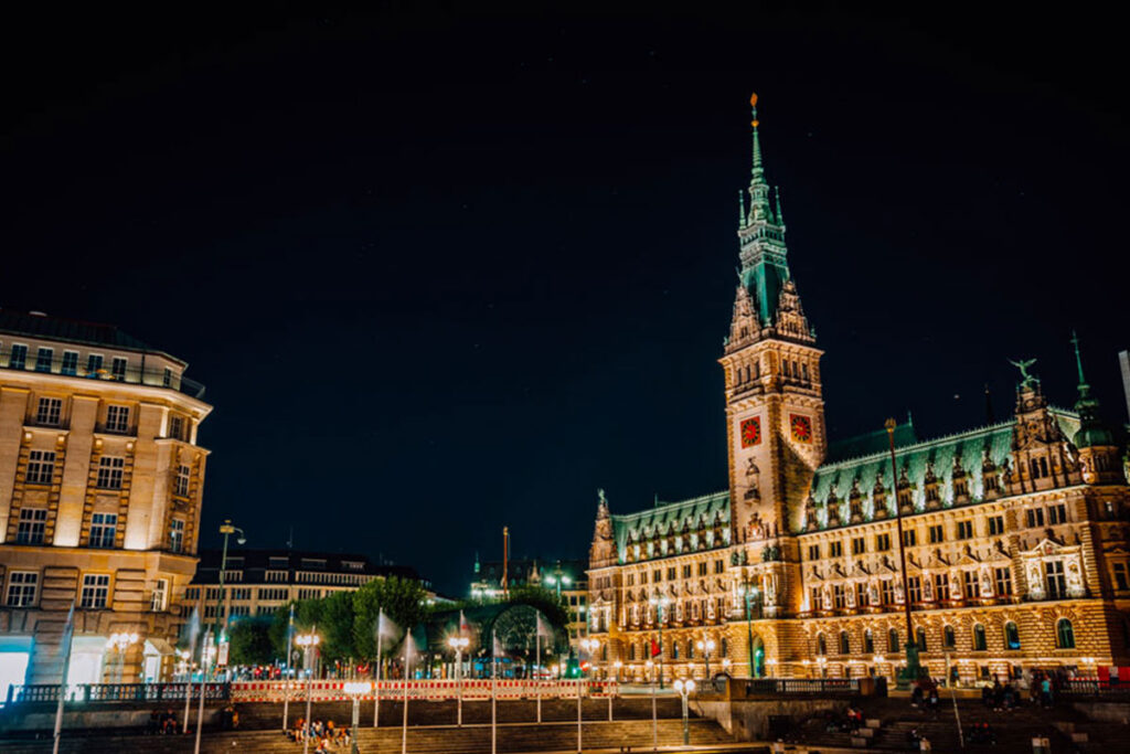 Beleuchtetes Gebäude im gotischen Stil mit einem hohen Uhrturm in Stuttgart bei Nacht, umgeben von beleuchteten Straßenlaternen und einem klaren, dunklen Himmel.