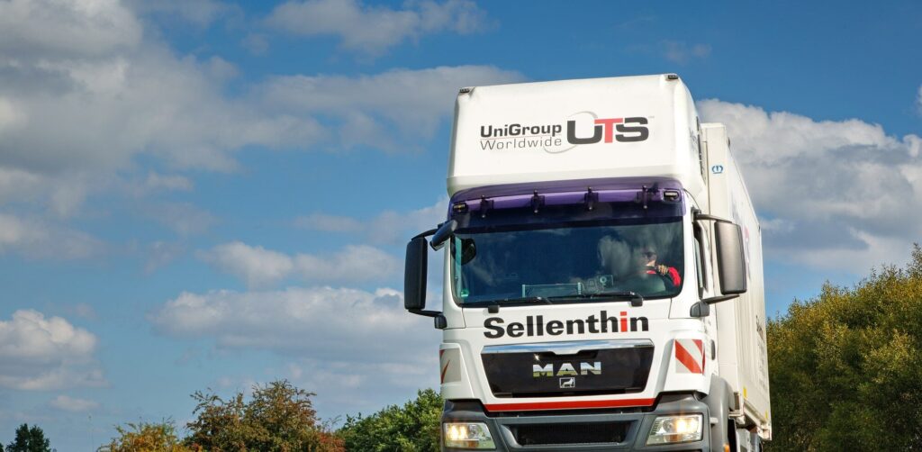 Weißer Lastwagen mit der Aufschrift „Sellenthin“ fährt auf einer Straße, umgeben von Bäumen, unter blauem Himmel mit Wolken.