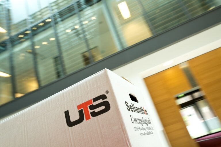 Ein Umzugskarton aus Pappe mit der Aufschrift „UTS“ in schwarzen und roten Buchstaben steht in einem modernen Gebäude. Der Hintergrund ist leicht unscharf und vermittelt Bewegung.